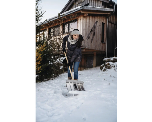 Frau schaufelt Schnee vor einem Holzhaus mit einer Schneeschaufel