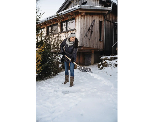 Frau schippt Schnee vor einem Holzhaus mit einer Schneeschaufel.