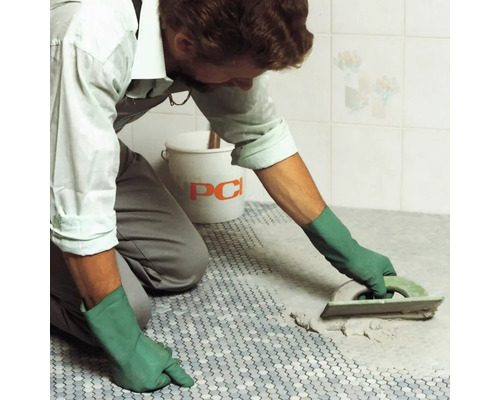 Fliesenleger verfugt Mosaikfliesen mit einem Fugenbrett im Badezimmer. Im Hintergrund befindet sich ein Eimer der Marke PCI.