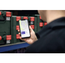 Eine Person hält ein Smartphone in der Hand, das eine Ortungs-App anzeigt. Im Hintergrund sind Werkzeugkoffer zu sehen.
