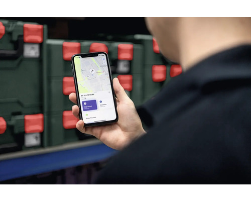 Eine Person hält ein Smartphone in der Hand, das eine Ortungs-App anzeigt. Im Hintergrund sind Werkzeugkoffer zu sehen.