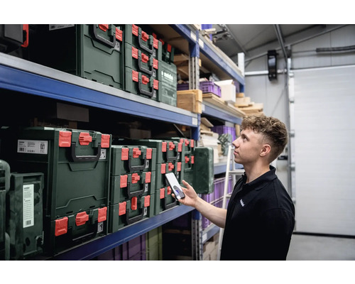 Regale mit Metabo Werkzeugkoffern in einem Lager, ein Mann inspiziert sie mit einem Mobiltelefon.