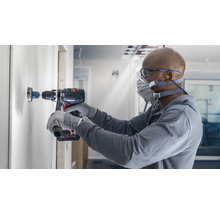 Handwerker bohrt mit einer Lochsäge ein Loch in eine Wand. Er trägt dabei eine Schutzmaske und Schutzbrille. Eine Bohrmaschine des Herstellers Bosch ist im Einsatz.