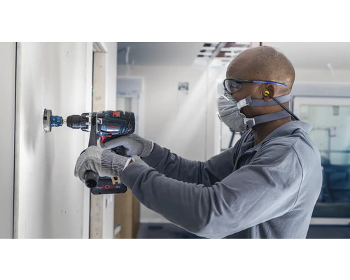 Handwerker bohrt mit einer Lochsäge ein Loch in eine Wand. Er trägt dabei eine Schutzmaske und Schutzbrille. Eine Bohrmaschine des Herstellers Bosch ist im Einsatz.
