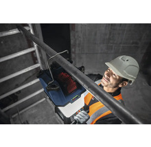 Arbeiter mit Schutzhelm befestigt eine Akku-Baustellenleuchte an einem Rohr.