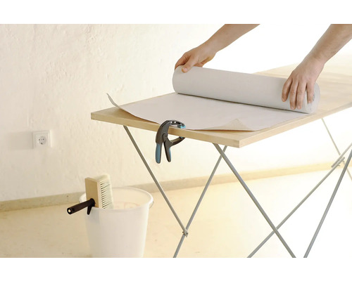 Person rollt Tapete auf einem Tapeziertisch aus. Eimer und Quast stehen bereit.