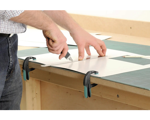 Zuschneiden einer Kunststoffplatte mit einem Cuttermesser und einem Lineal, fixiert mit einer Zwinge.