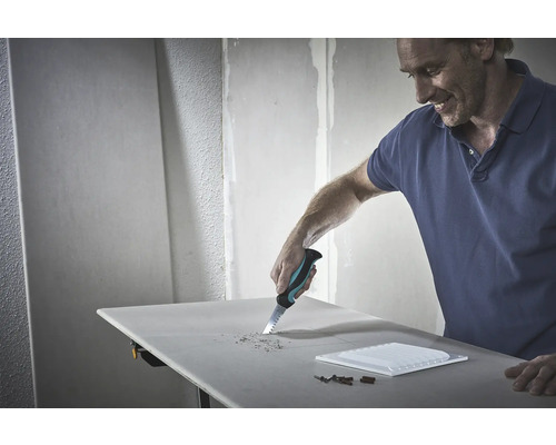 Ein Mann sägt mit einer Handsäge ein Loch in eine Gipskartonplatte. Werkzeuge und eine Fliese liegen auf dem Tisch.