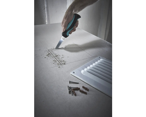 Handwerker sägt mit einer Gipskartonsäge ein Loch in eine Gipskartonplatte, daneben ein Lüftungsgitter sowie Schrauben und Dübel