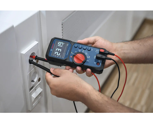 Ein Mann misst mit einem Digitalmultimeter die Spannung an einer Steckdose.
