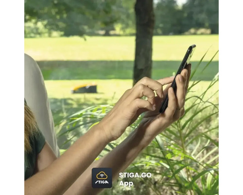 Hände bedienen die Stiga Go App auf einem Smartphone, Rasenmähroboter im Hintergrund