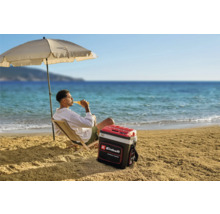 Ein Mann entspannt am Strand unter einem Sonnenschirm mit einer Einhell Kühlbox neben sich