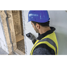 Mann mit Bauhelm und Schutzweste benutzt Laser-Entfernungsmesser an einer Wand.