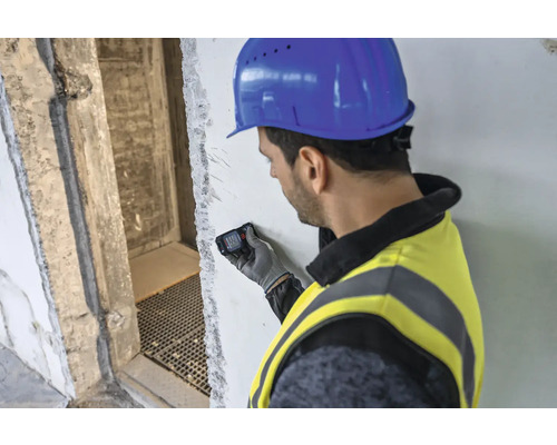 Mann mit Bauhelm und Schutzweste benutzt Laser-Entfernungsmesser an einer Wand.