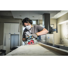 Eine Person benutzt eine Kreissäge zum Schneiden eines Holzbretts, geschützt mit Maske, Brille und Gehörschutz.