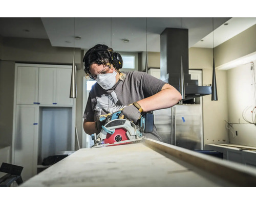 Eine Person benutzt eine Kreissäge zum Schneiden eines Holzbretts, geschützt mit Maske, Brille und Gehörschutz.