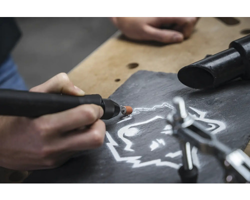 Nahaufnahme einer Person, die mit einem Multifunktionswerkzeug ein Wolfsmotiv in einen Schieferstein graviert.