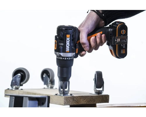Eine Hand hält einen Akkuschrauber der Marke Worx beim Anbringen einer Lenkrolle an einem Holzbrett.