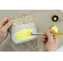 Eine Person bemalt einen Pflanzkorb mit einem Pinsel und gelber Farbe.