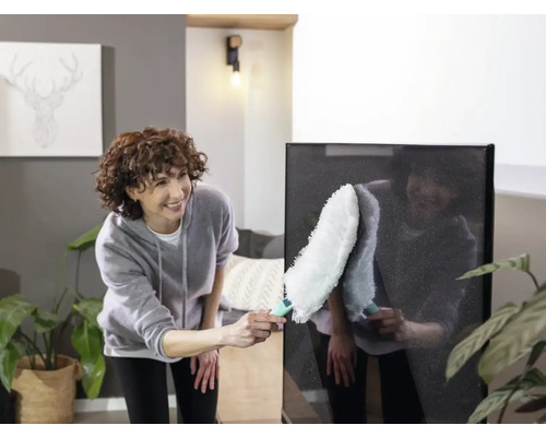 Frau reinigt einen Flachbildfernseher mit einem Staubwedel in einem hellen Wohnzimmer.