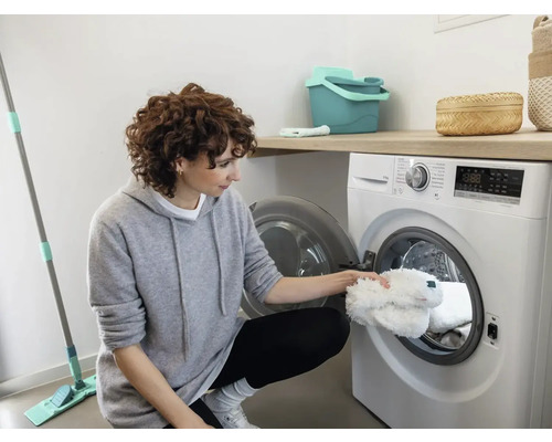 Frau legt weißen Moppbezug in eine Waschmaschine in einem hellen Raum. Reinigungseimer und Wischmopp sind ebenfalls sichtbar.