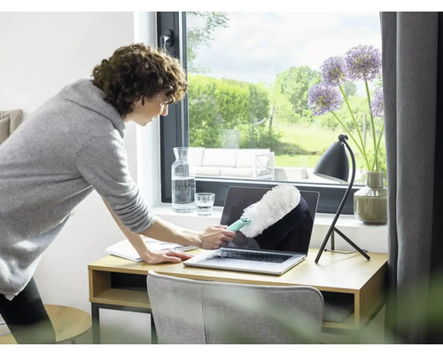Frau reinigt Laptop mit Staubwedel auf Schreibtisch.