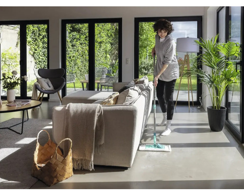 Frau wischt Boden in hellem Wohnzimmer mit großen Fenstern und minimalistischer Einrichtung.