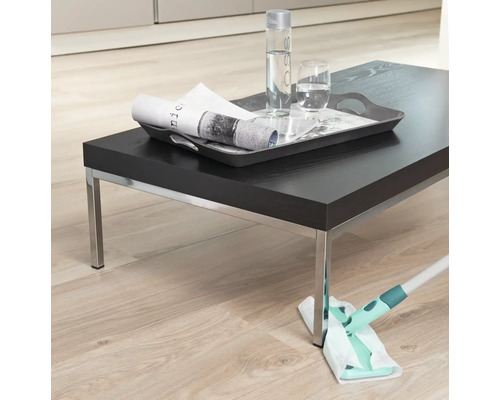 Szene mit einer Person, die mit einem Mopp einen Holzboden reinigt, auf dem ein dunkler Tisch mit Getränken steht.