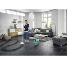 Frau wischt Fliesenboden mit Wischmopp und Eimer in einem hellen Wohnzimmer.