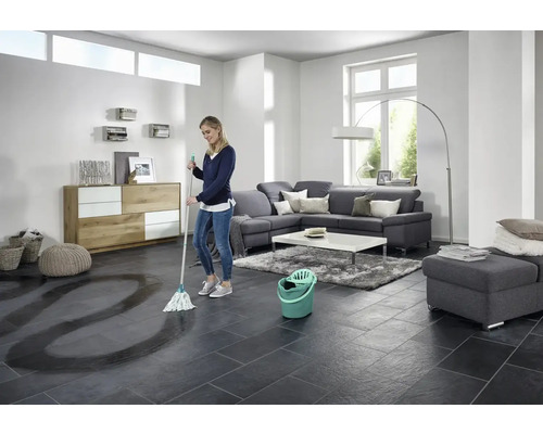 Frau wischt Fliesenboden mit Wischmopp und Eimer in einem hellen Wohnzimmer.