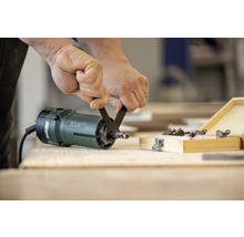 Person wechselt Fräser an Metabo Oberfräse mit Schraubenschlüssel neben Holzbox mit Fräser-Set.