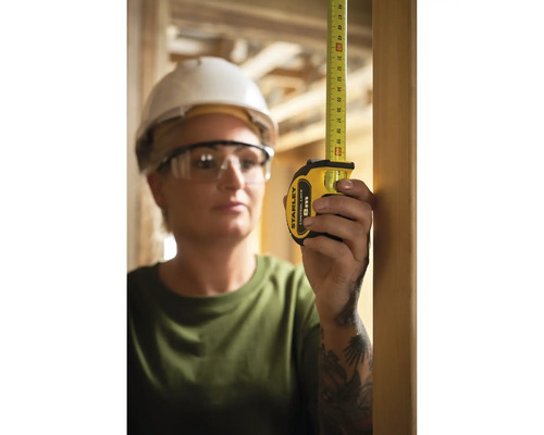 Frau mit Schutzbrille und Bauhelm misst mit einem Stanley Rollbandmaß.