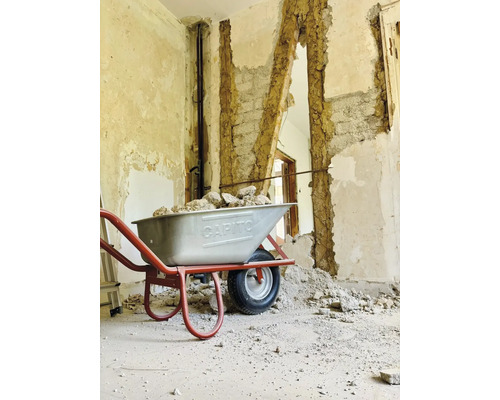 Eine Schubkarre gefüllt mit Bauschutt vor einer teilweise abgerissenen Wand.