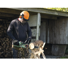 Mann sägt Holz mit einer elektrischen Kettensäge auf einem Holzbock, trägt dabei Schutzkleidung.
