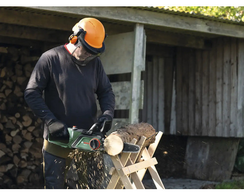 Mann sägt Holz mit einer elektrischen Kettensäge auf einem Holzbock, trägt dabei Schutzkleidung.