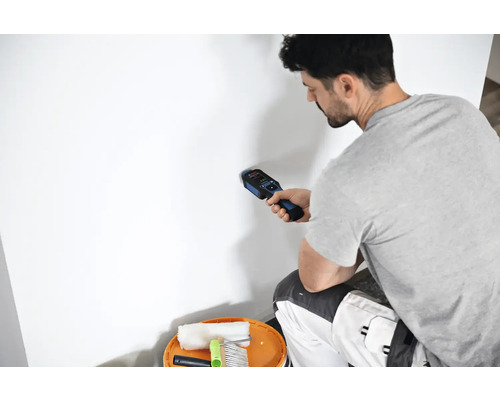 Mann benutzt einen Detektor an einer Wand in der Nähe eines Eimers mit Malerwerkzeug