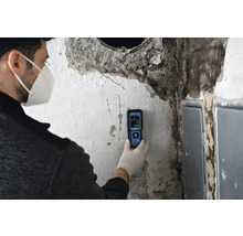 Eine Person mit Schutzmaske und Handschuhen führt mit einem Feuchtigkeitsmessgerät eine Feuchtigkeitsmessung an einer Wand durch.