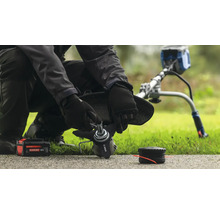 Eine Person wechselt die Klinge eines Rasentrimmers aus, wobei eine Batterie und eine Spule in der Nähe liegen.