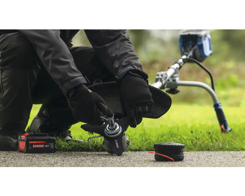 Eine Person wechselt die Klinge eines Rasentrimmers aus, wobei eine Batterie und eine Spule in der Nähe liegen.