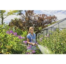 Frau gießt Blumen mit einem Gartenschlauch in einem grünen Garten vor einem Gewächshaus.
