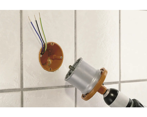 Diamant-Lochsäge bohrt Loch in geflieste Wand neben einer Verteilerdose mit elektrischen Kabeln.