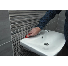 Eine Person entfernt Silikonfugen zwischen Waschbecken und Wandfliesen mit einem Werkzeug.