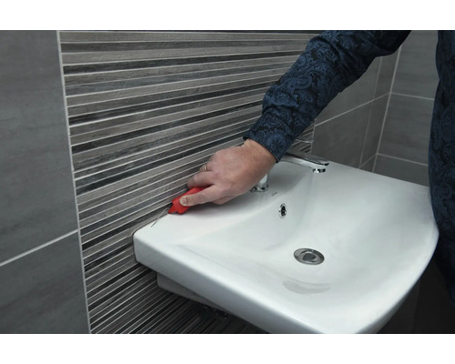 Eine Person entfernt Silikonfugen zwischen Waschbecken und Wandfliesen mit einem Werkzeug.