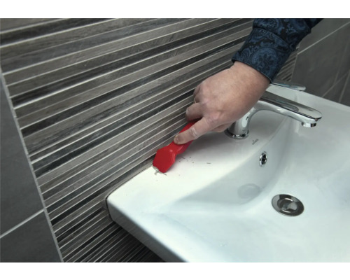 Eine Person entfernt Dichtungsmasse mit einem Fugenkratzer von einem Waschbecken