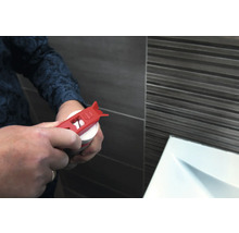 Eine Person öffnet eine Kartusche mit einem roten Kartuschenöffner vor einer gefliesten Wand.