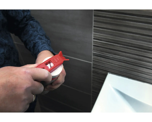 Eine Person öffnet eine Kartusche mit einem roten Kartuschenöffner vor einer gefliesten Wand.