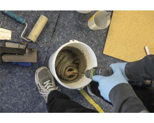 Mischen von Fliesenkleber im Eimer mit einem elektrischen Rührer. Werkzeuge und eine Person mit Schutzhandschuhen sind ebenfalls zu sehen.