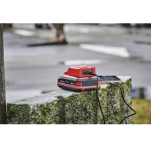 Ein Einhell Power X-Change Akku mit USB-Adapter lädt ein Smartphone auf einer Steinmauer.