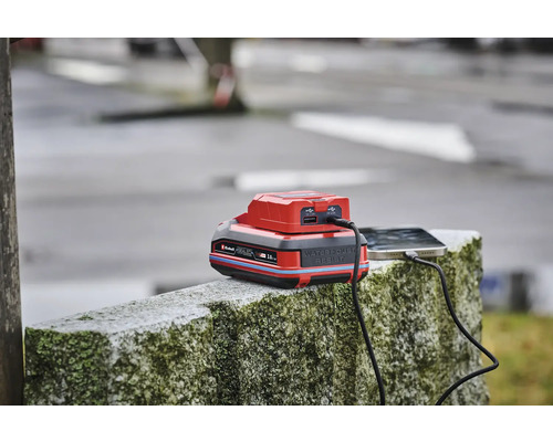 Ein Einhell Power X-Change Akku mit USB-Adapter lädt ein Smartphone auf einer Steinmauer.