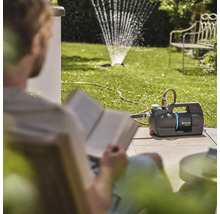 Garten mit Rasensprenger und lesendem Mann neben einer Gartenpumpe